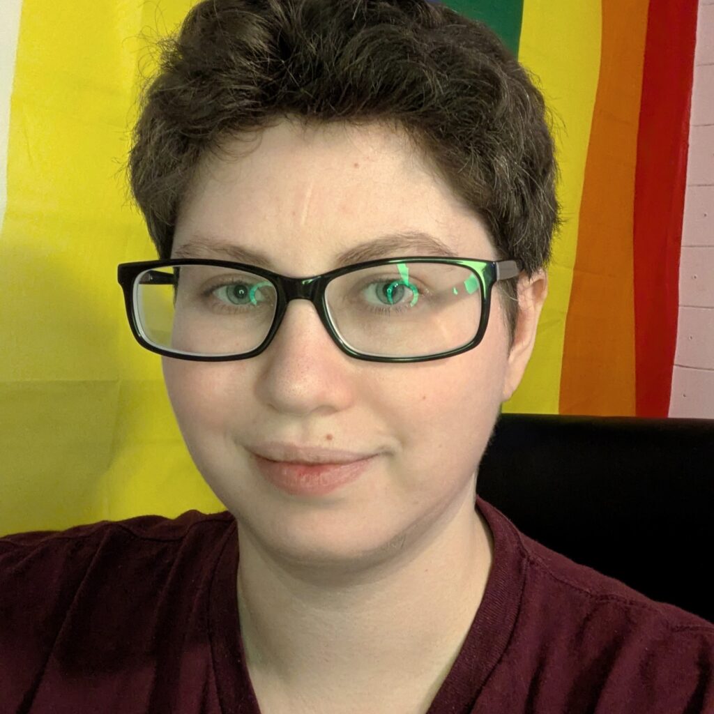 A person with pale skin, dark brown hair, and blue eyes, with black glasses and a burgundy t-shirt. They have a yellow, orange, and red background.